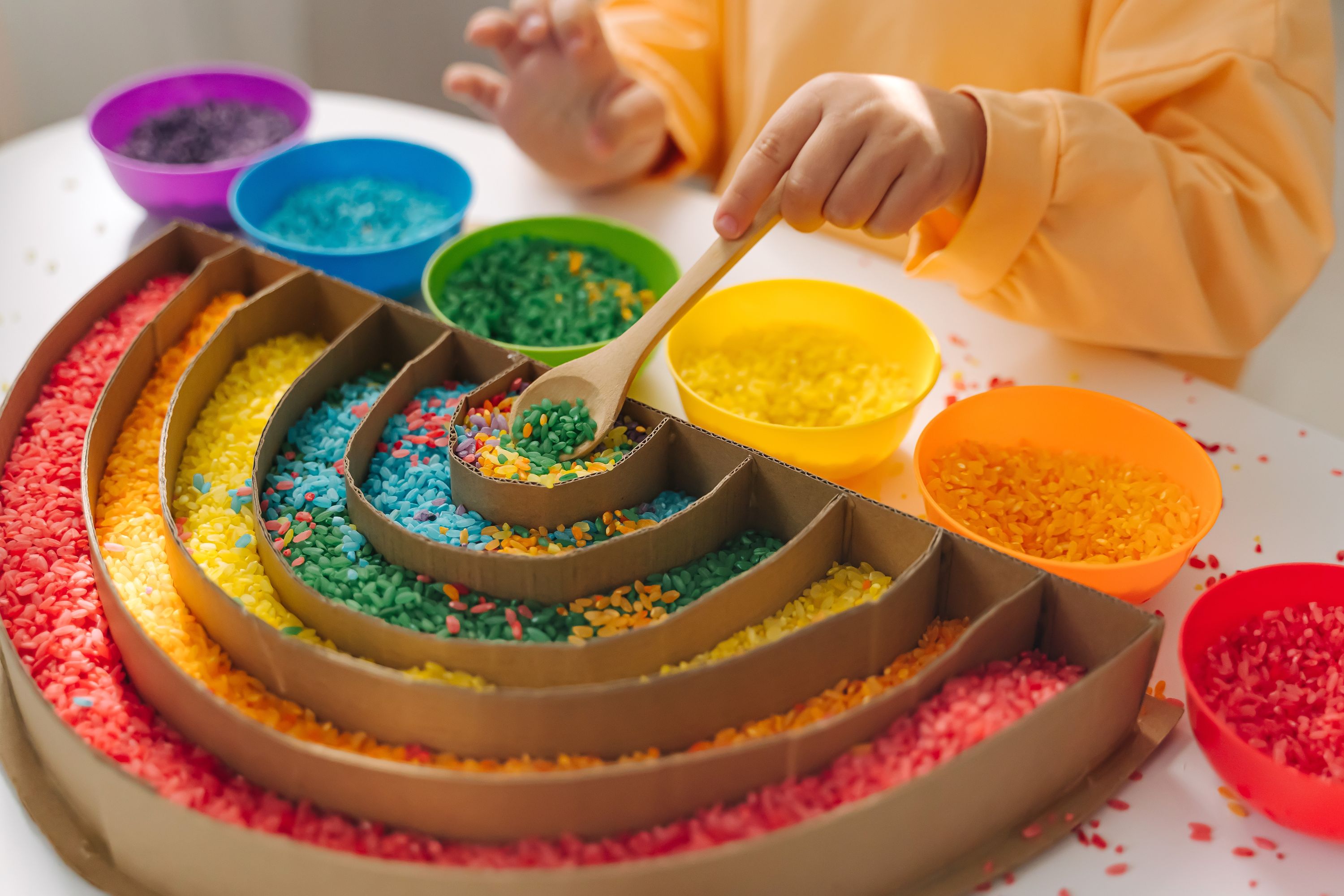 Child filling cardboard rainbow with coloured rice