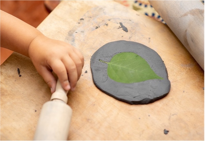 Child with rolling pin next to leaf pressed in clay 