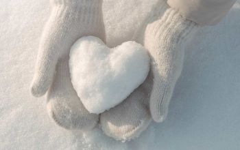 Hands wearing beige mittens holding snow heart against snow background