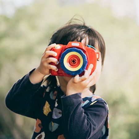 Child looking through a toy camera with an unidentifiable outdoor background.